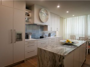 Custom kitchen cabinets Sarasota — contemporary white flat-panel cabinetry with quartzite waterfall island, blue tile backsplash, and integrated refrigerator by EuroTech Cabinetry